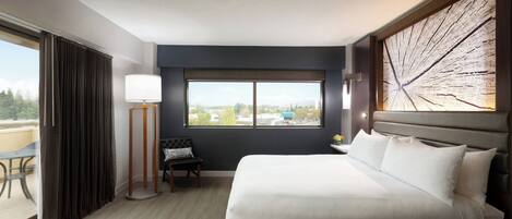 Habitación de hotel con una cama grande, vista a la ciudad por una ventana, un banco y una lámpara de pie.