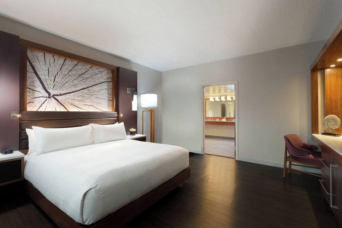 Ein modernes Hotelzimmer mit einem großen Bett, einem Schreibtisch mit Stuhl, einem Nachttisch, einer Wandlampe und einem Spiegel.