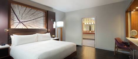 A modern hotel room with a large bed, a desk with a chair, a nightstand, a wall-mounted lamp, and a mirror.