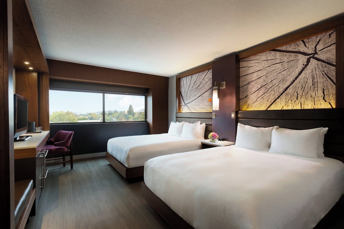 Ein Hotelzimmer mit zwei Betten, einem Schreibtisch mit Stuhl, einem Fernseher und einem großen Fenster.