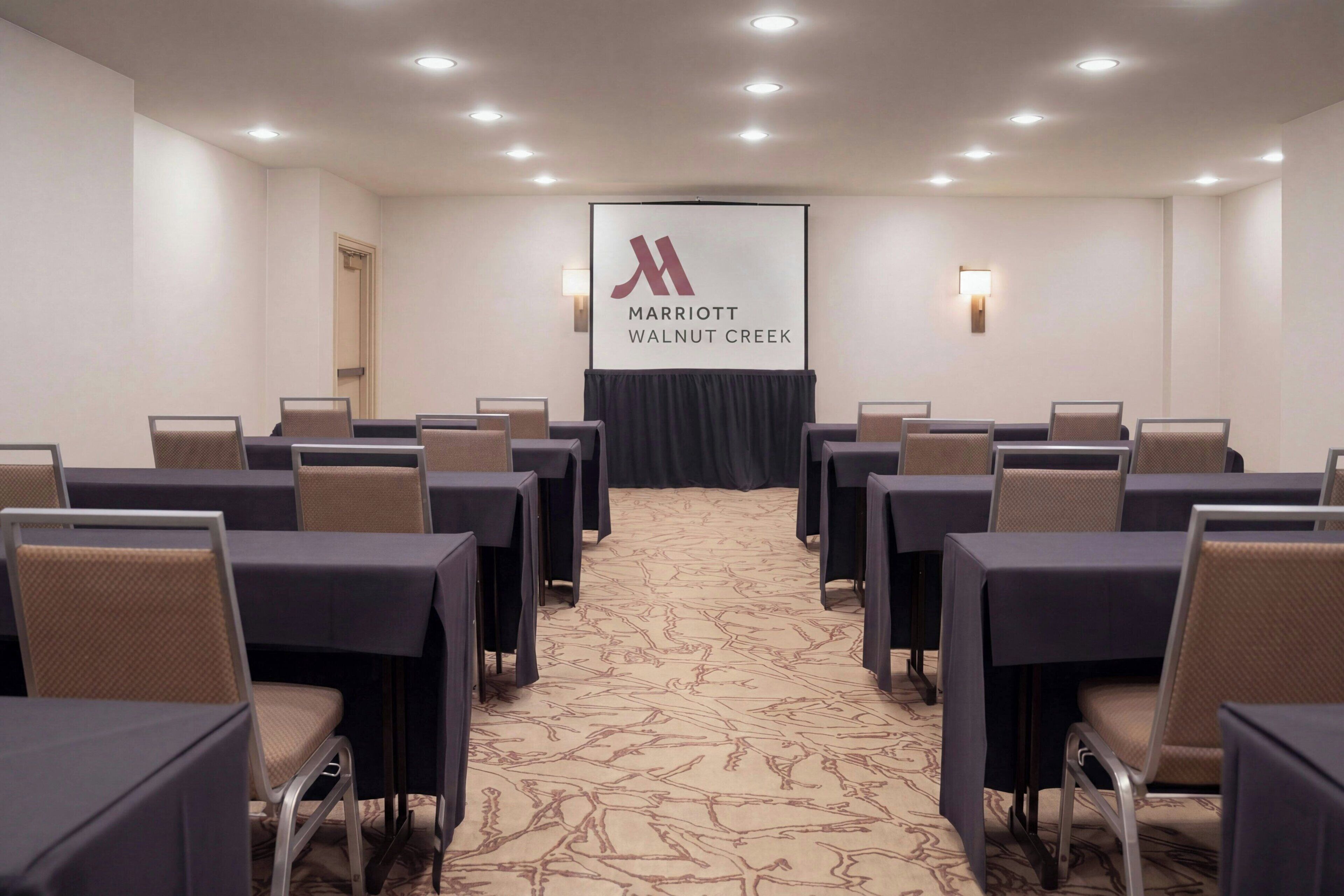 A conference room with a large screen displaying the Marriott logo, rows of tables and chairs, and recessed ceiling lighting.