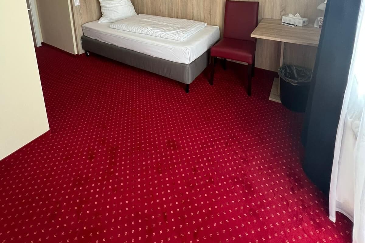 Une chambre d’hôtel avec un lit, un bureau, une chaise, un tableau au mur et une moquette rouge.