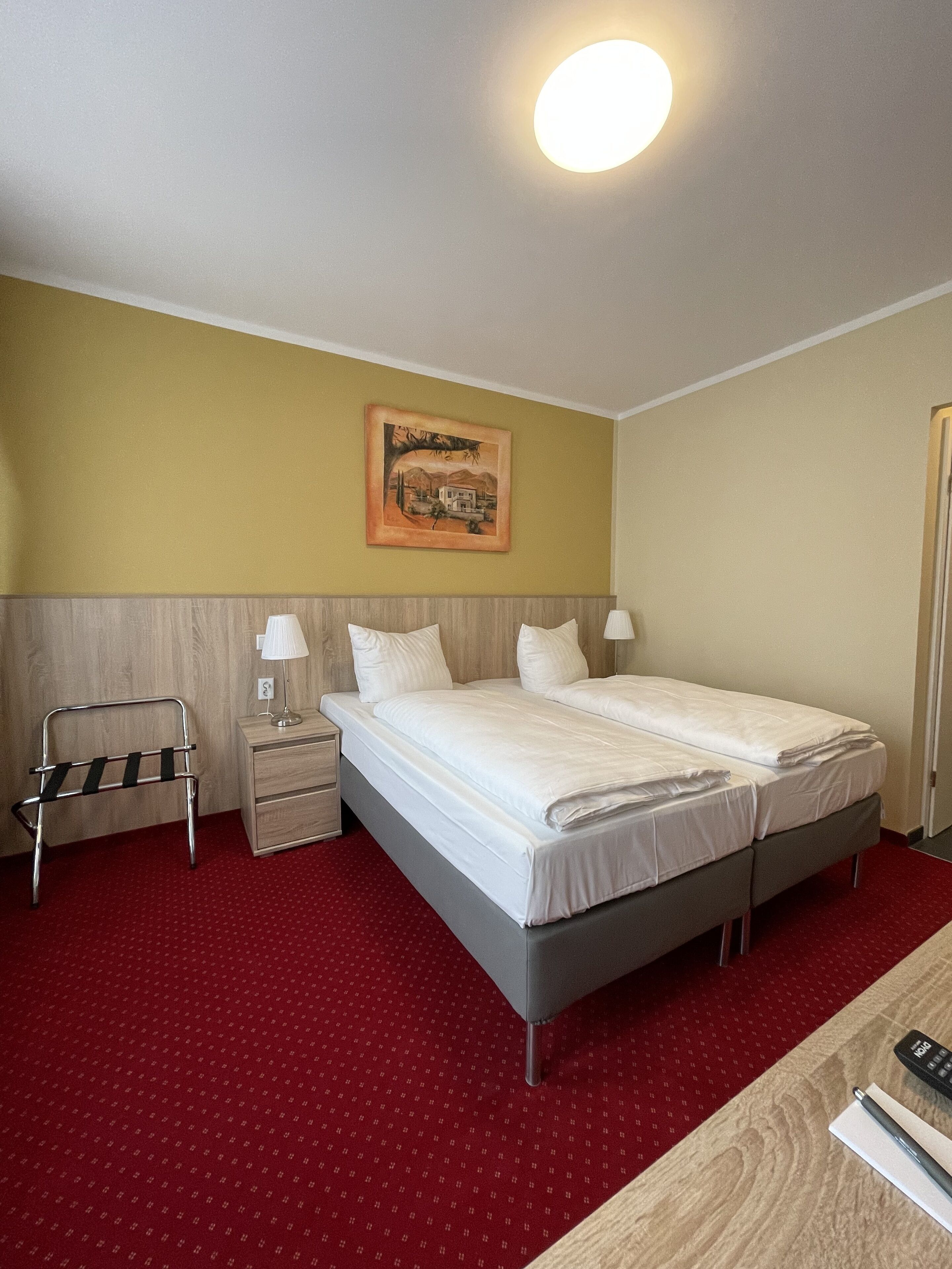A hotel room with a bed, bedside tables, a lamp, a painting on the wall, and a red carpet.