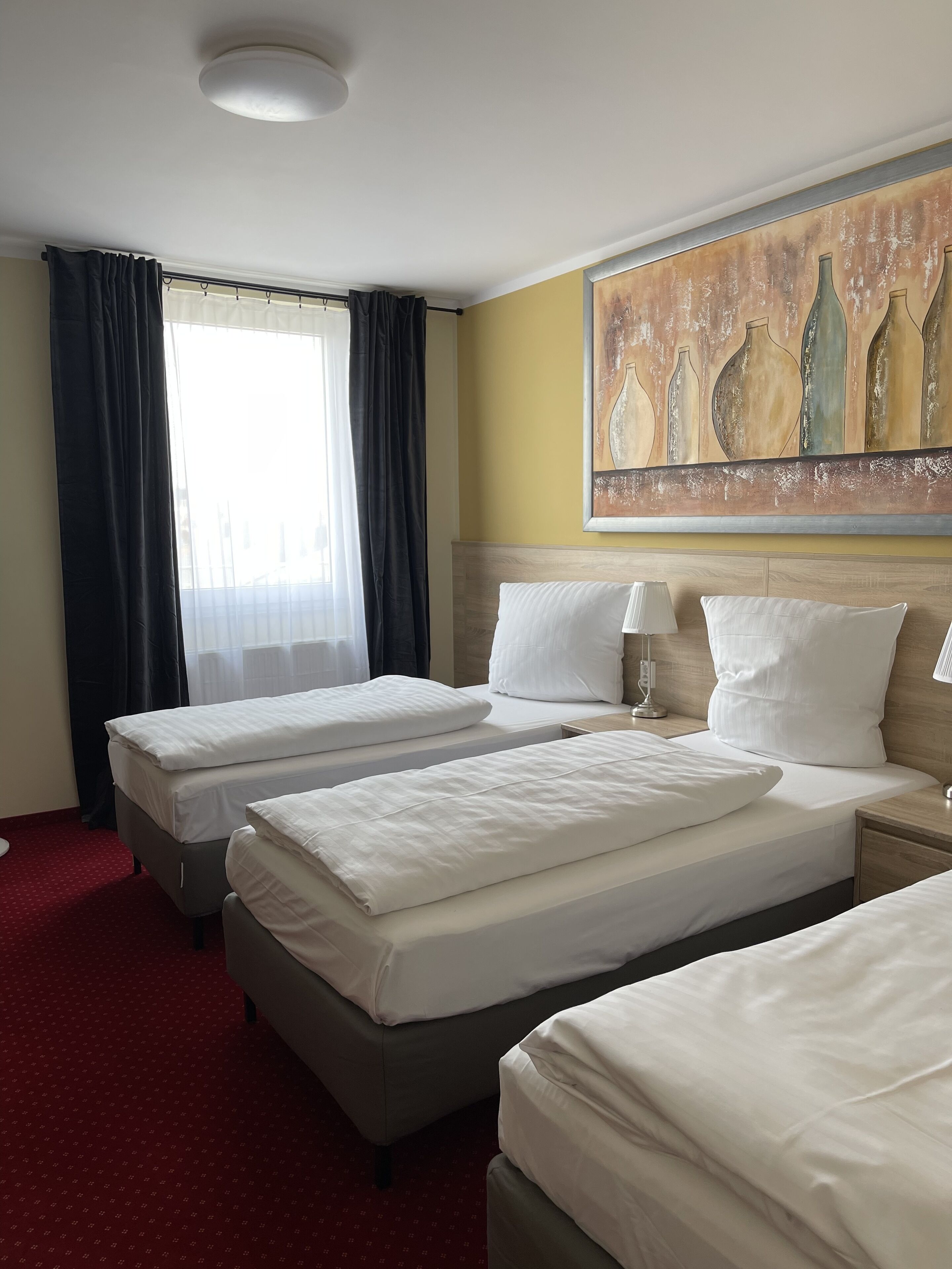 Ein Hotelzimmer mit zwei Betten, einem großen Fenster mit Vorhängen, einem Wandbild und einem roten Teppich.
