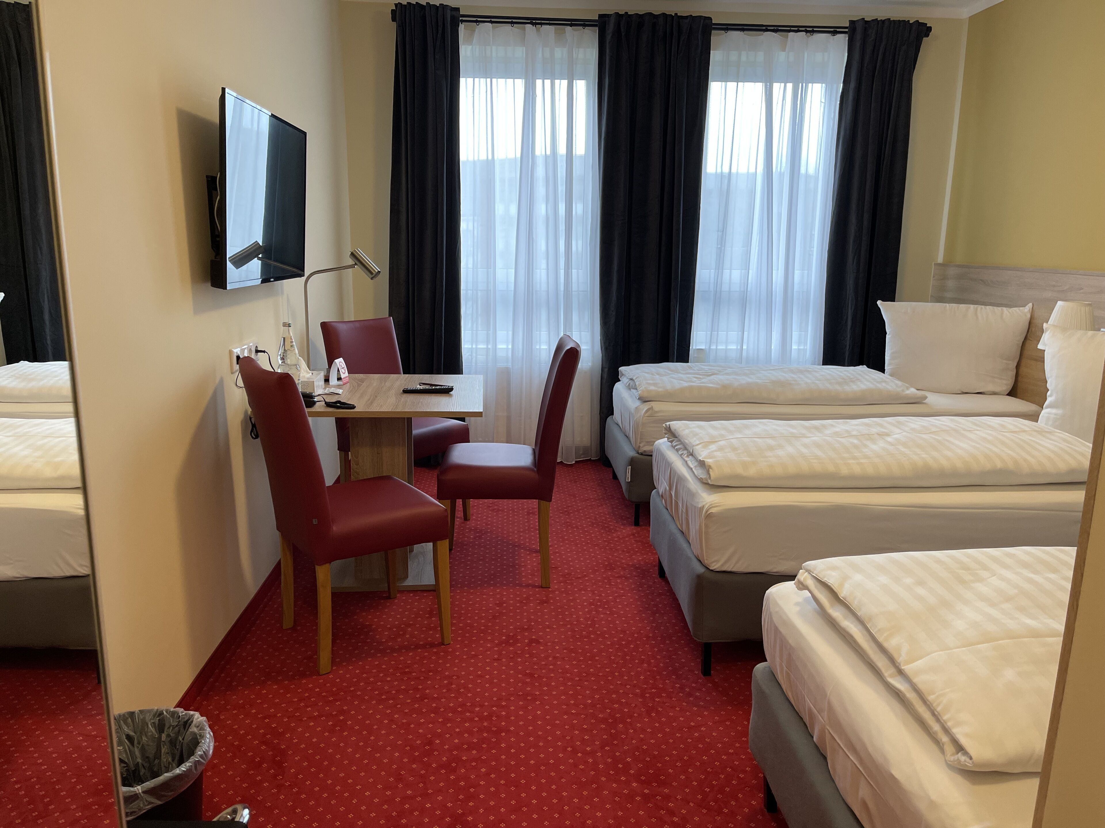 A hotel room with a bed, a desk with a chair, a TV mounted on the wall, and a window with curtains.