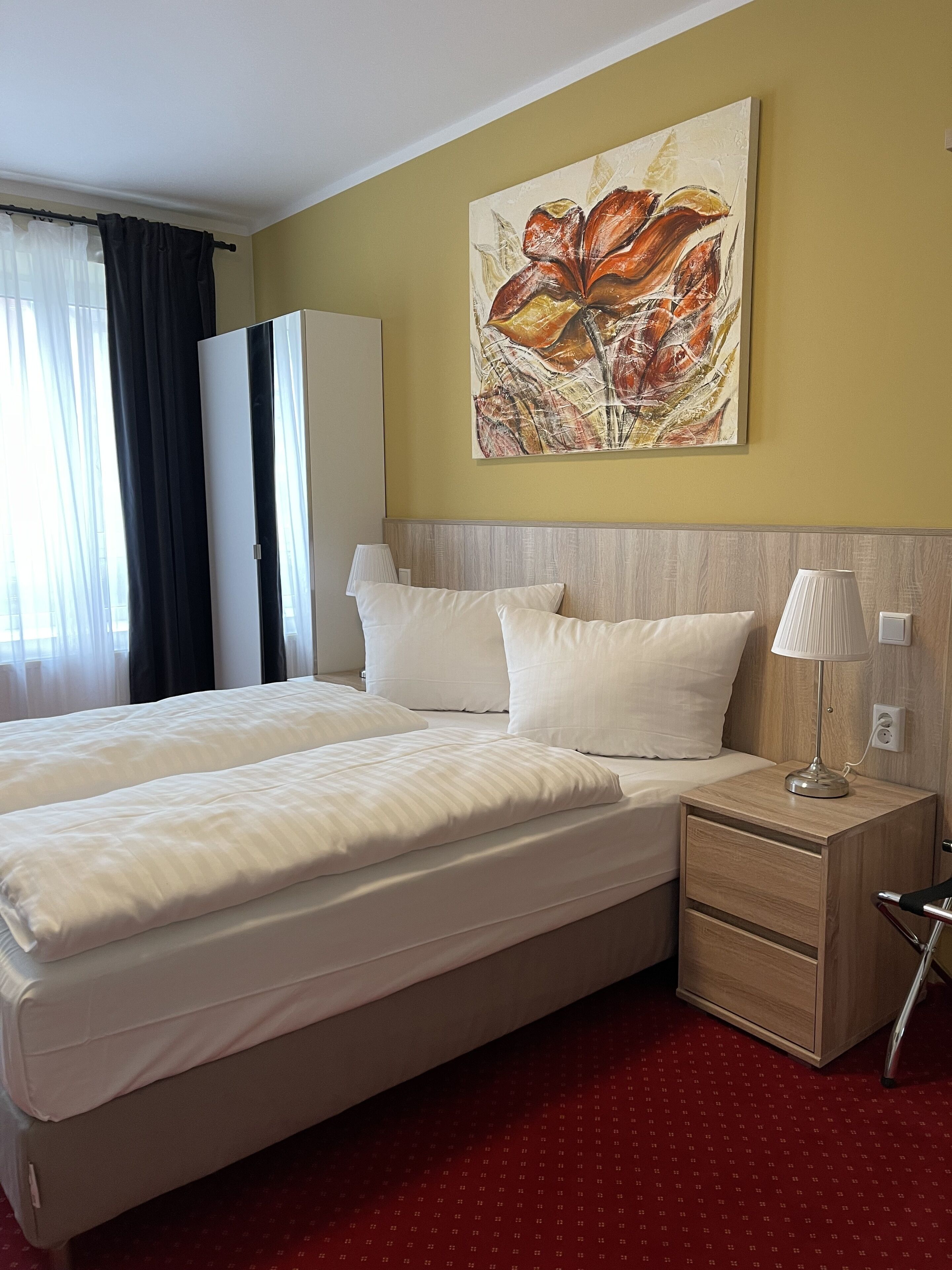 Una habitación de hotel con una cama grande, una mesita de noche con lámpara, un armario y un cuadro en la pared.