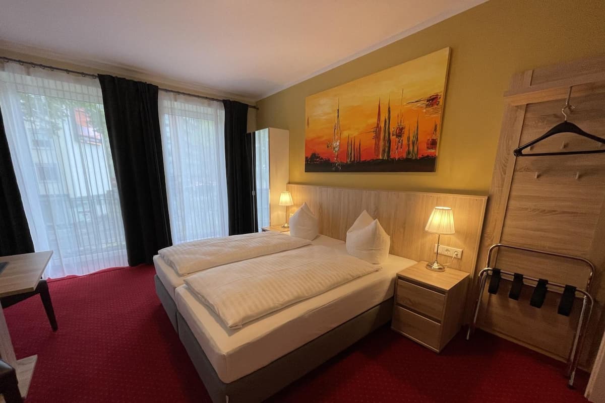 Une chambre d’hôtel avec un lit double, deux tables de chevet avec des lampes, une armoire, un bureau et une grande fenêtre avec des rideaux.
