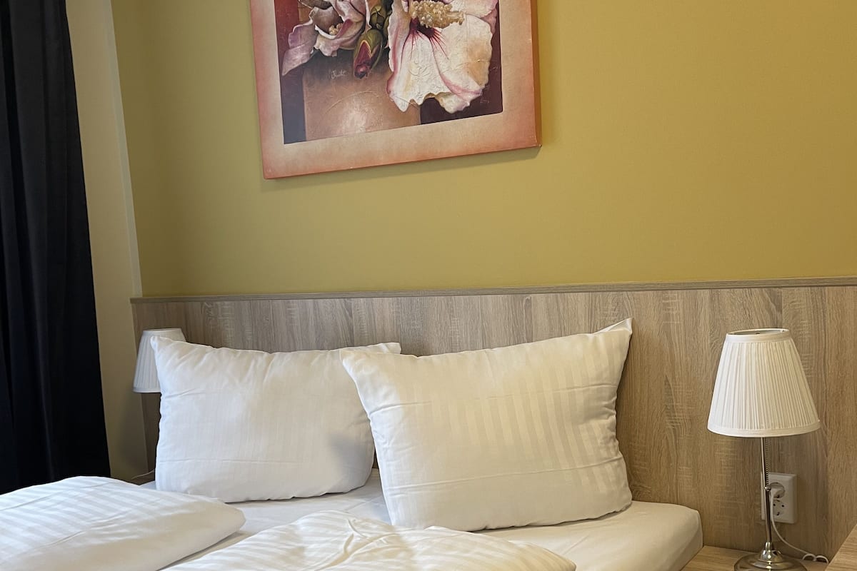Une chambre d’hôtel avec un lit, deux oreillers, une table de chevet avec une lampe et un tableau représentant des fleurs accroché au mur.
