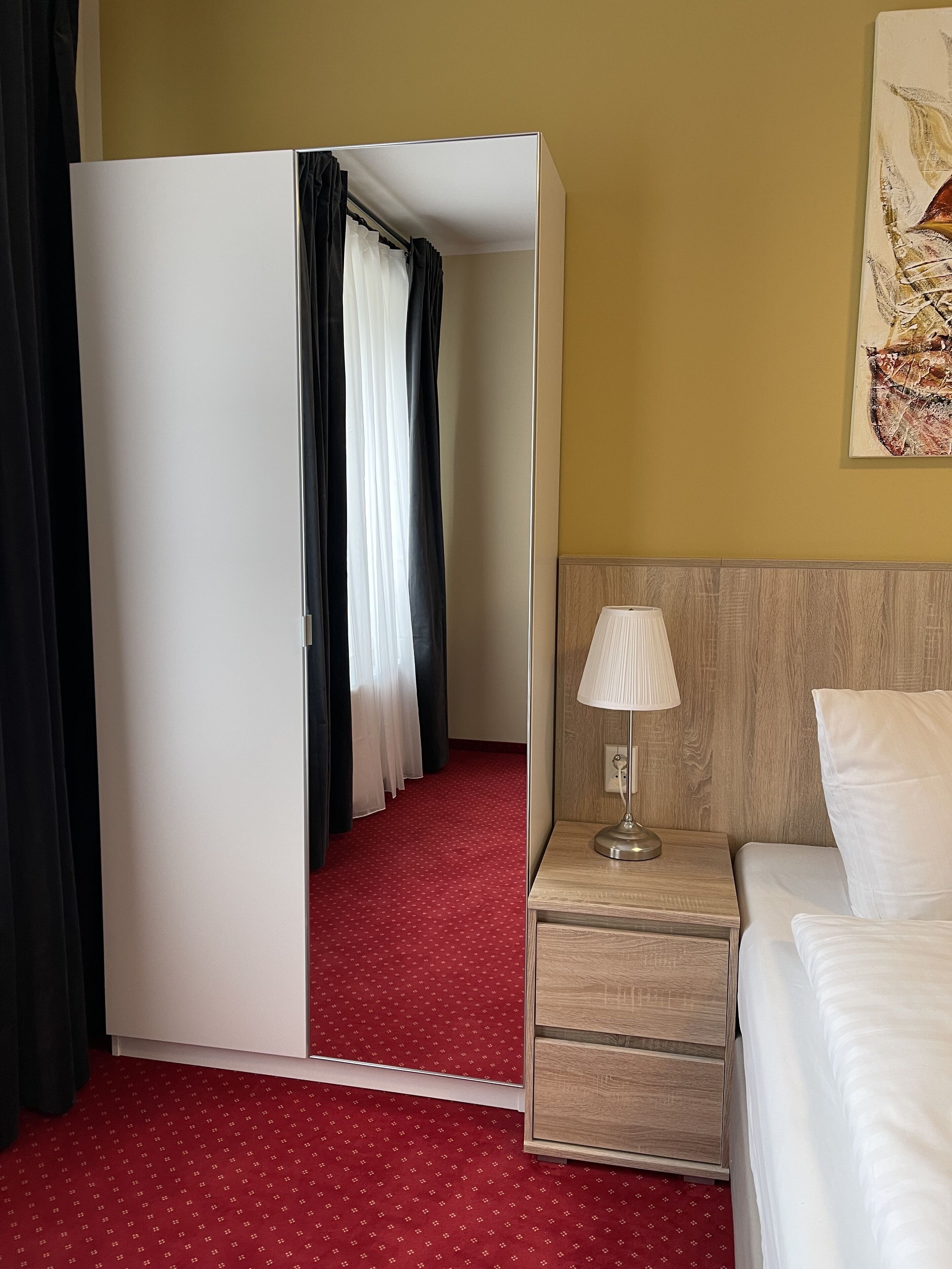 Habitación de hotel con cama, mesita de noche con lámpara, cabecera de madera, alfombra roja, armario y espejo.
