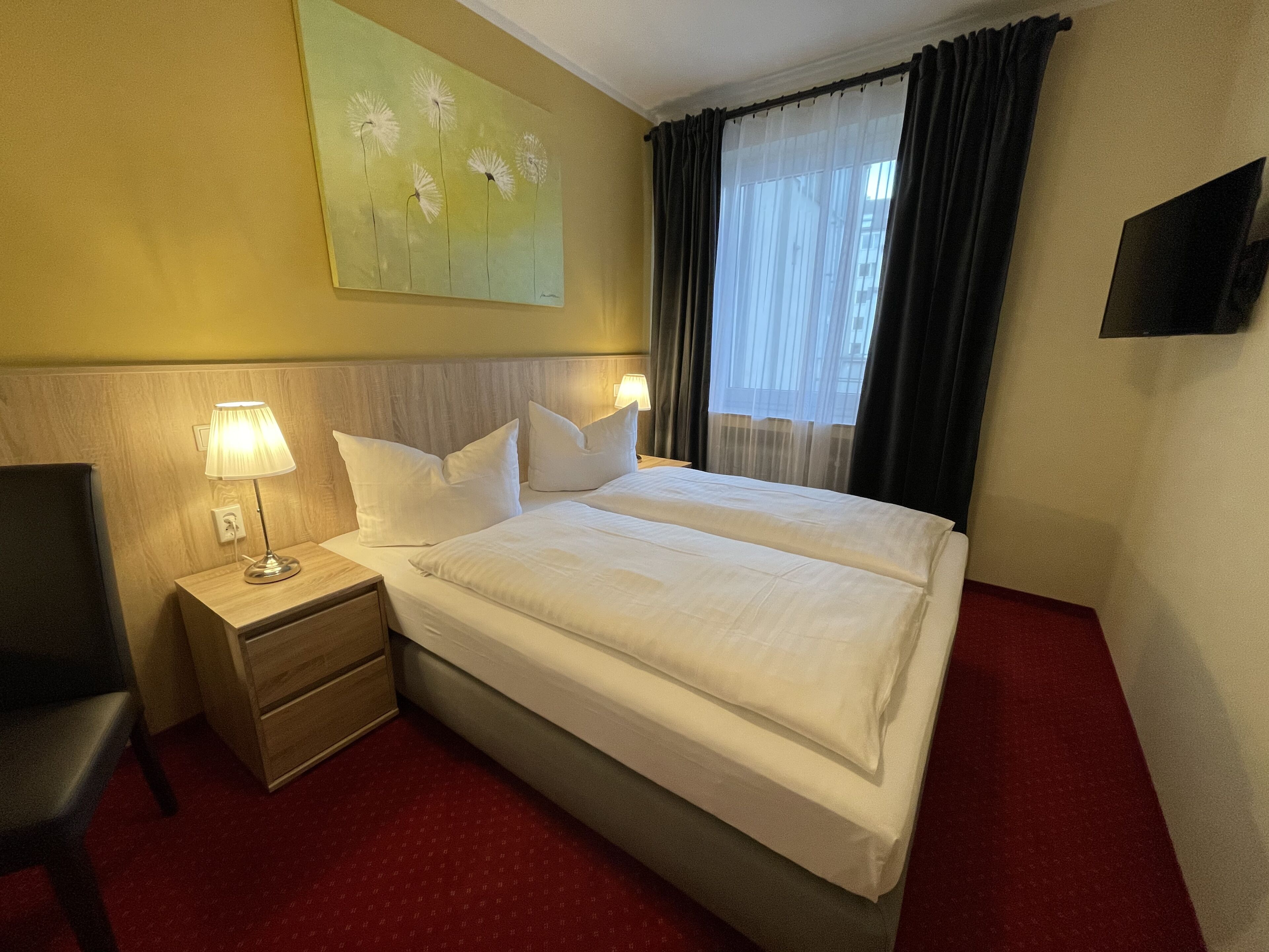 Un cuarto de hotel con una cama grande, dos lámparas de noche, una mesita de noche de madera, una silla, un cuadro en la pared y un televisor montado en la pared.