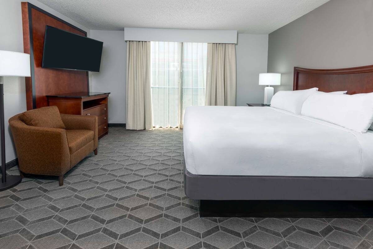 Una habitación de hotel con una cama grande, un televisor montado en la pared, un escritorio con una silla y una ventana con cortinas.