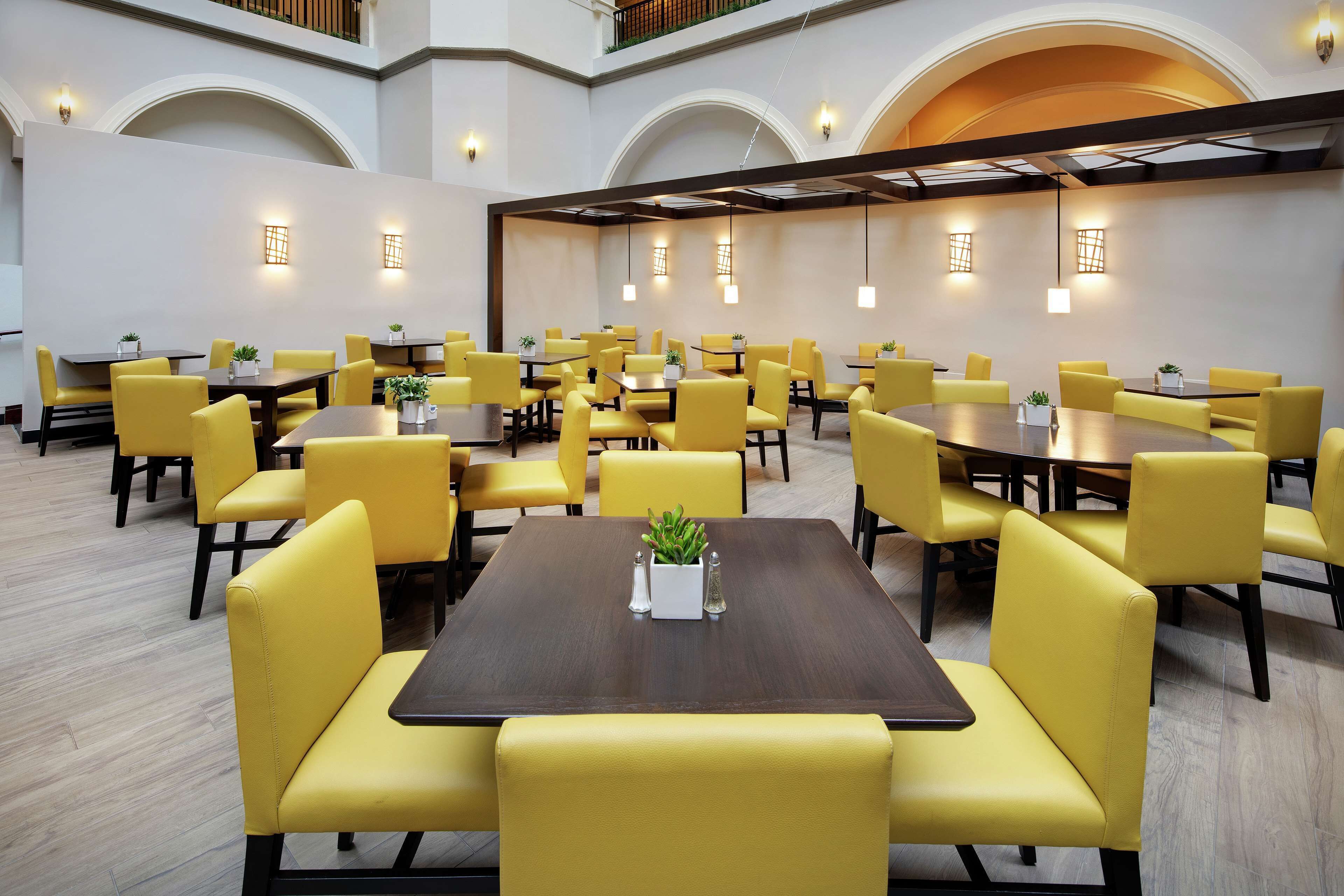A spacious dining area with yellow chairs and tables, featuring a central plant and salt and pepper shakers.