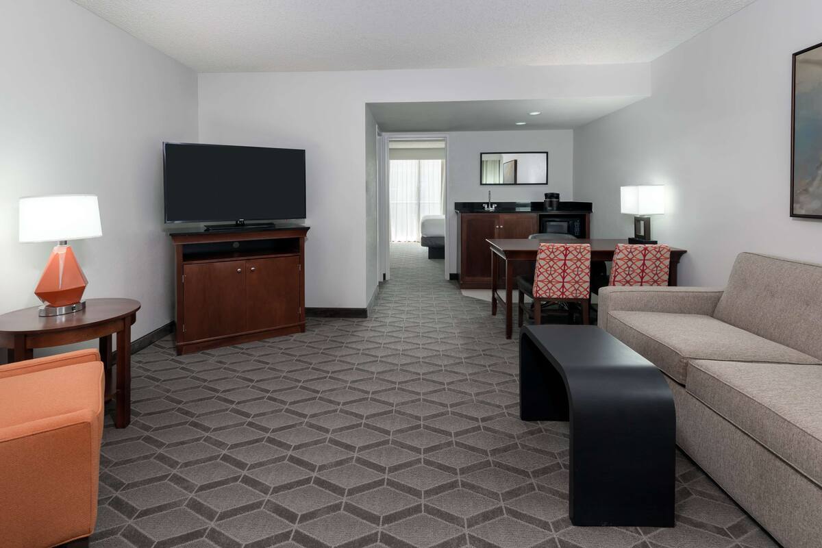 Habitación de hotel con televisor, sofá, mesa de comedor con sillas, zona de cocina y pasillo que conduce a otra habitación.