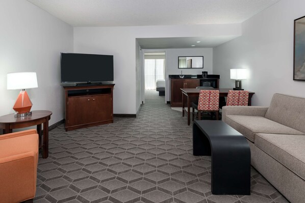 A hotel room with a TV, a sofa, a dining table with chairs, a kitchen area, and a hallway leading to another room.