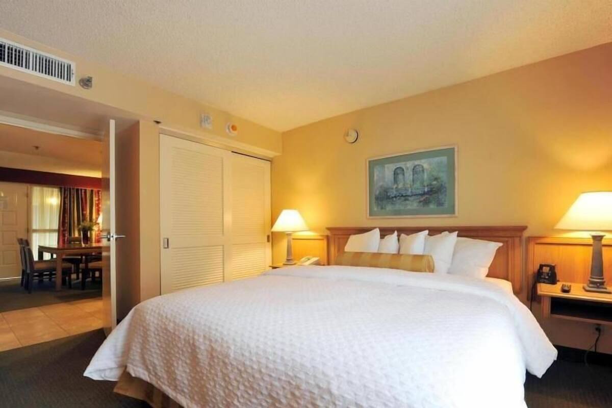 Habitación de hotel con una cama grande, dos lámparas de noche, un cuadro en la pared y una puerta que da a un comedor.