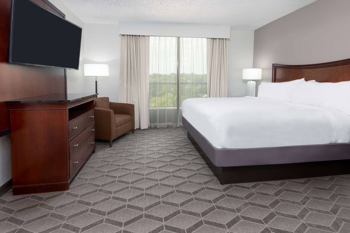 Una habitación de hotel con una cama grande, un televisor montado en la pared, un tocador, una silla y una ventana con cortinas.