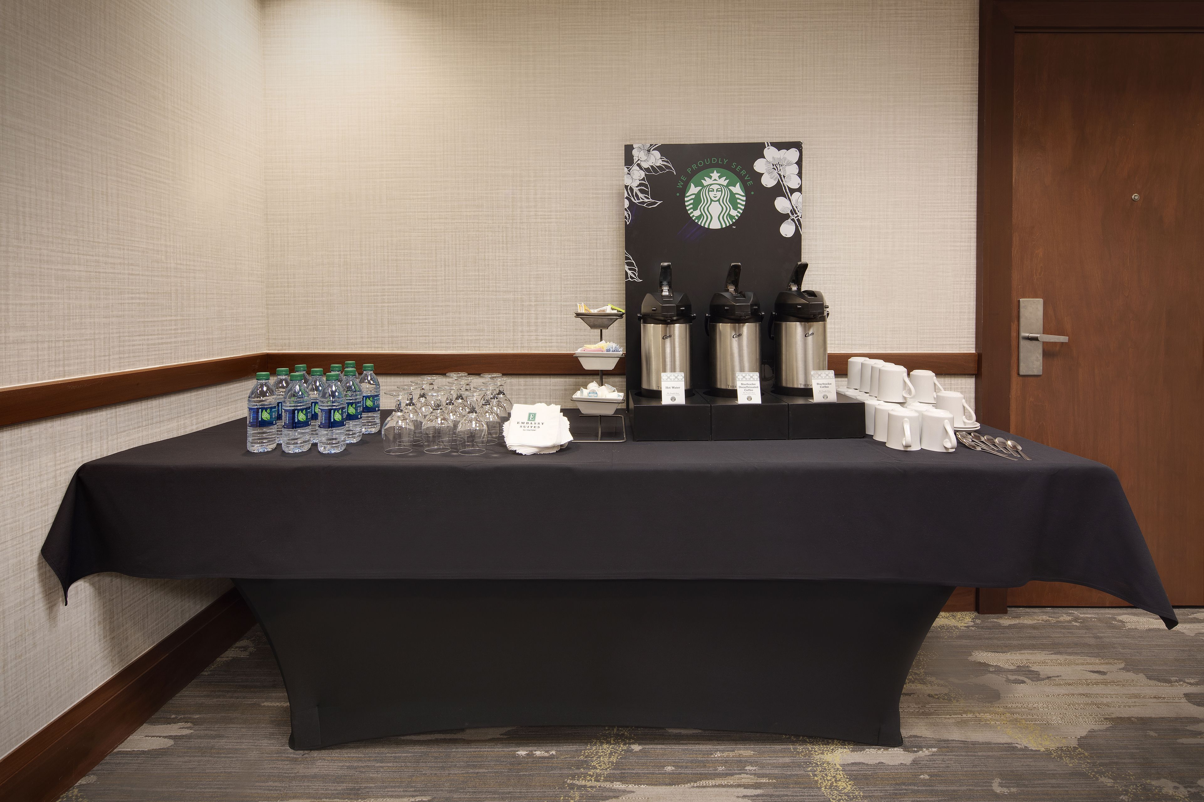 Ein Tisch mit Flaschenwasser, Gläsern und Kaffeemaschinen, vor einer Wand mit floralem Dekor und dem Starbucks-Logo.
