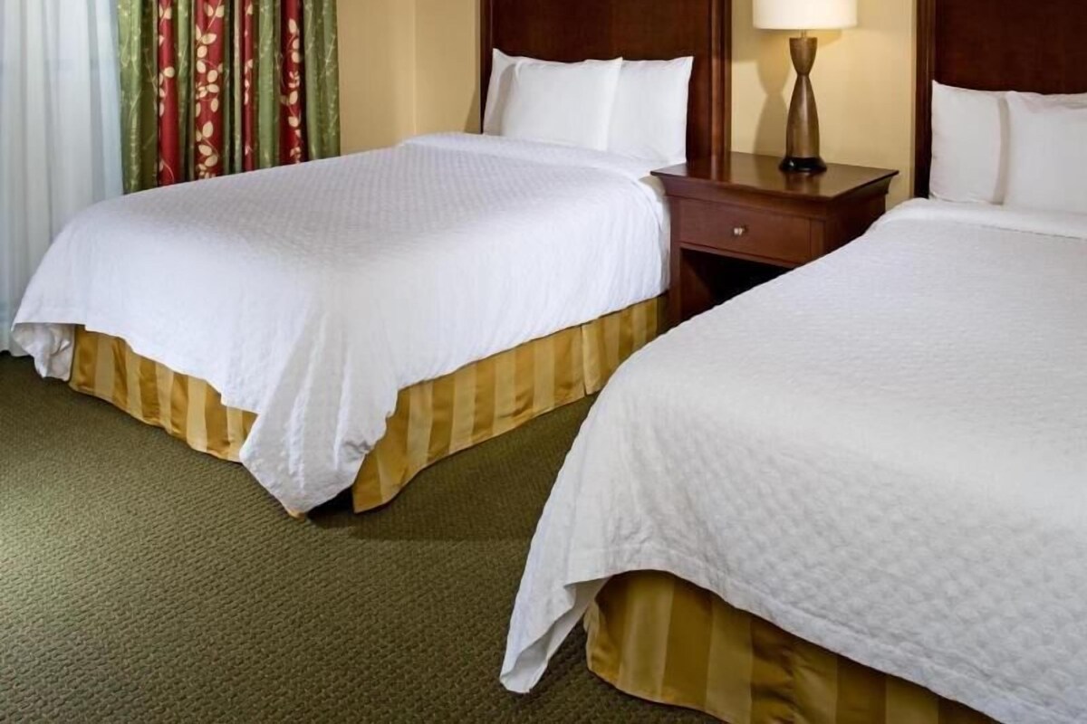 Une chambre d’hôtel avec deux lits, une table de chevet avec une lampe et une fenêtre avec des rideaux.