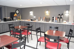Una cafetería moderna con paredes grises, paneles de techo blancos y una variedad de dispensadores de comida y bebida. Hay varias mesas y sillas, y un reloj en la pared.