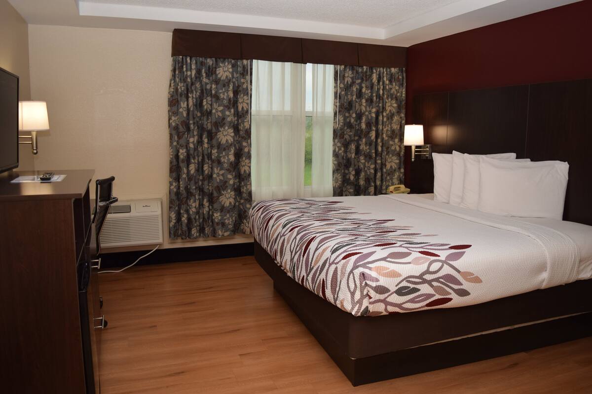 Une chambre d’hôtel avec un grand lit, un bureau avec une lampe, une télévision et une fenêtre avec des rideaux.