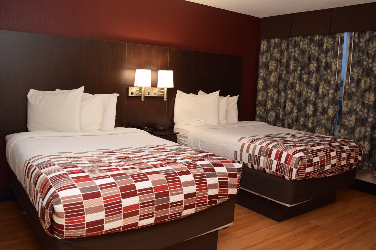 A hotel room with two beds, a nightstand, wall-mounted lamps, and a floral curtain.