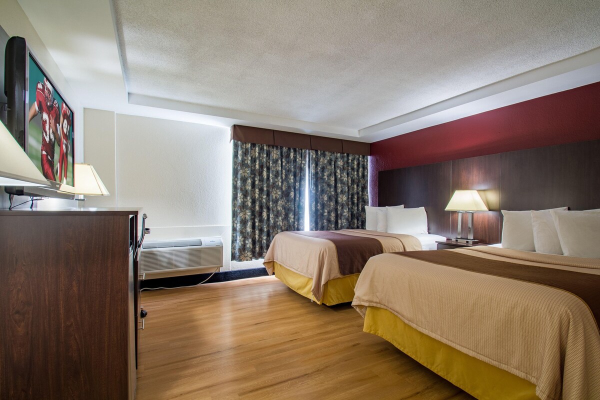 A hotel room with two beds, a desk with a lamp, a flat-screen TV, and a window with curtains.