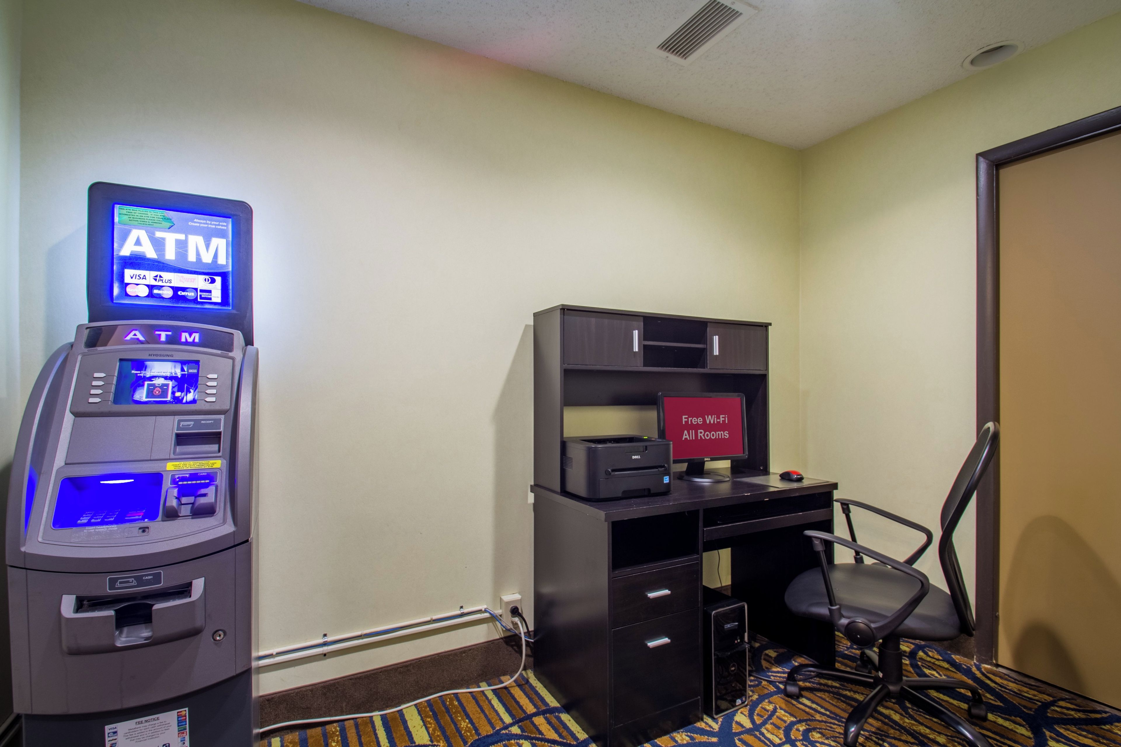 Una habitación de hotel con un cajero automático, un escritorio con una computadora y una silla.