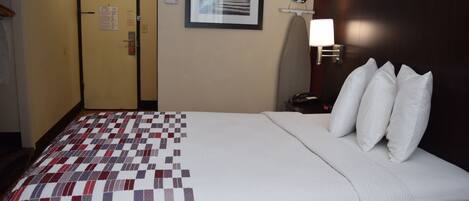 A hotel room with a bed, a quilt, a nightstand with a lamp, a framed picture on the wall, and a door with a sign.