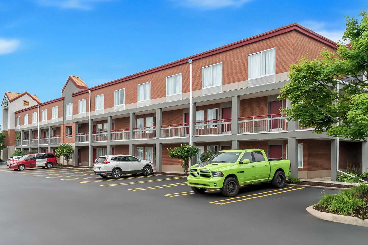 A multi-story motel with a parking lot, featuring a green pickup truck, a red car, and a white SUV.