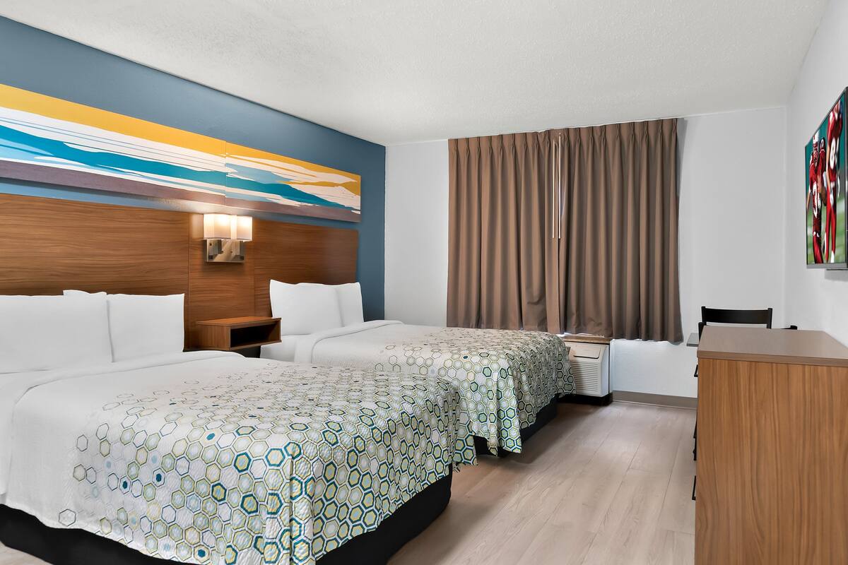 A hotel room with two beds, a desk, a chair, a wall-mounted lamp, and a large abstract painting.