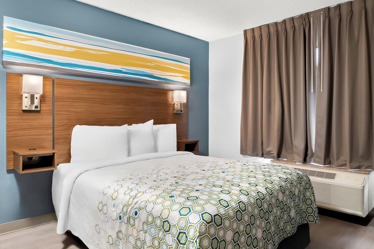 A hotel room with a bed, a headboard with a light fixture, a wall-mounted air conditioner, and a window with curtains.