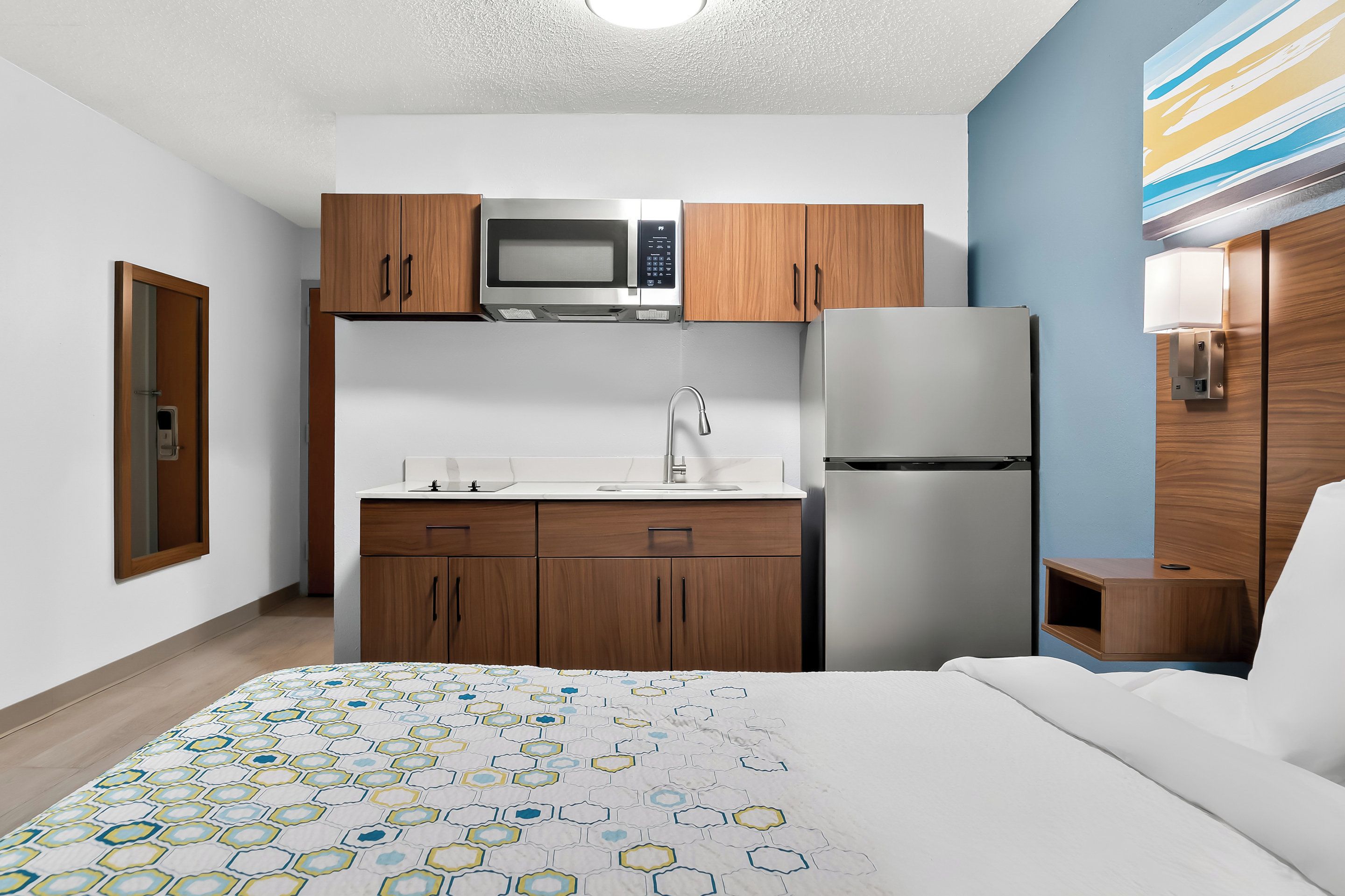 A compact hotel room with a kitchenette, a bed with a patterned bedspread, and a wooden nightstand.