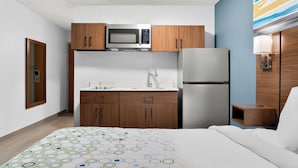 A compact hotel room with a kitchenette, a bed with a patterned bedspread, and a wooden nightstand. - HomeTowne Studios by Red Roof Auburn Hills (Auburn Hills)