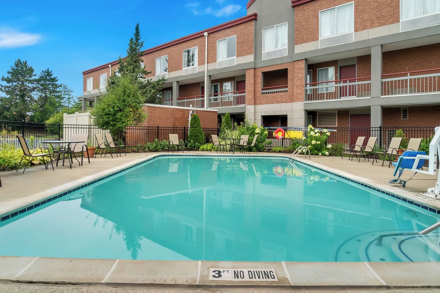 A rectangular pool with a 3-foot depth limit sign, surrounded by lounge chairs and a brick building with balconies.