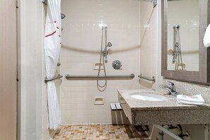 A bathroom with a shower, grab bars, a sink, and a mirror. - HomeTowne Studios by Red Roof Auburn Hills (Auburn Hills)