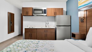 A compact hotel room with a kitchenette, a bed with a patterned bedspread, and a wooden nightstand. - HomeTowne Studios by Red Roof Auburn Hills (Auburn Hills)