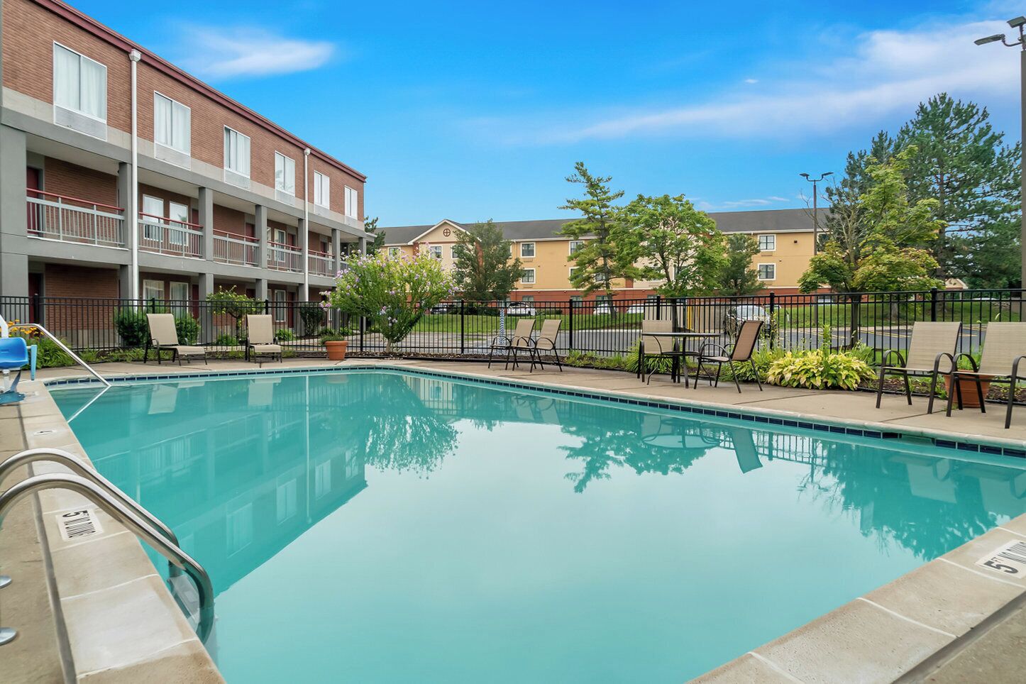 A swimming pool with lounge chairs and a fence, surrounded by a building with balconies and a parking area.