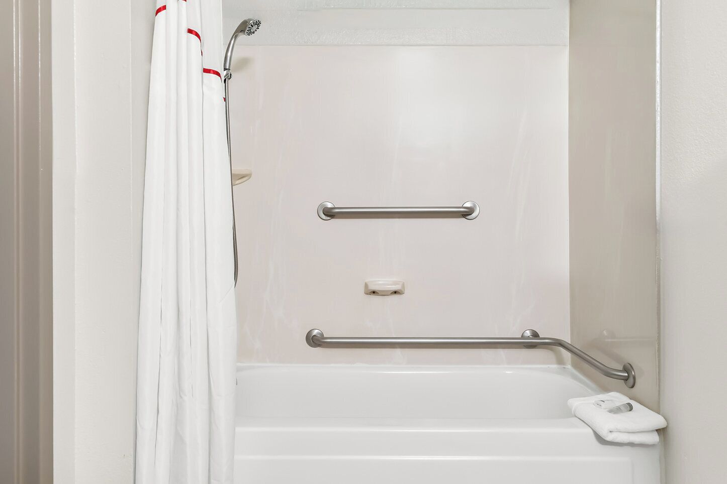 Baño blanco con ducha, barras de apoyo y cabezal de ducha manual.