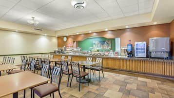 A breakfast area with tables, chairs, a counter with food and beverages, and a clock on the wall.