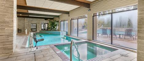 An indoor swimming pool with clear glass walls, wooden beams, and tiled flooring.