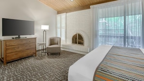 A bedroom with a bed, a TV on a dresser, a chair, a fireplace, and large windows.