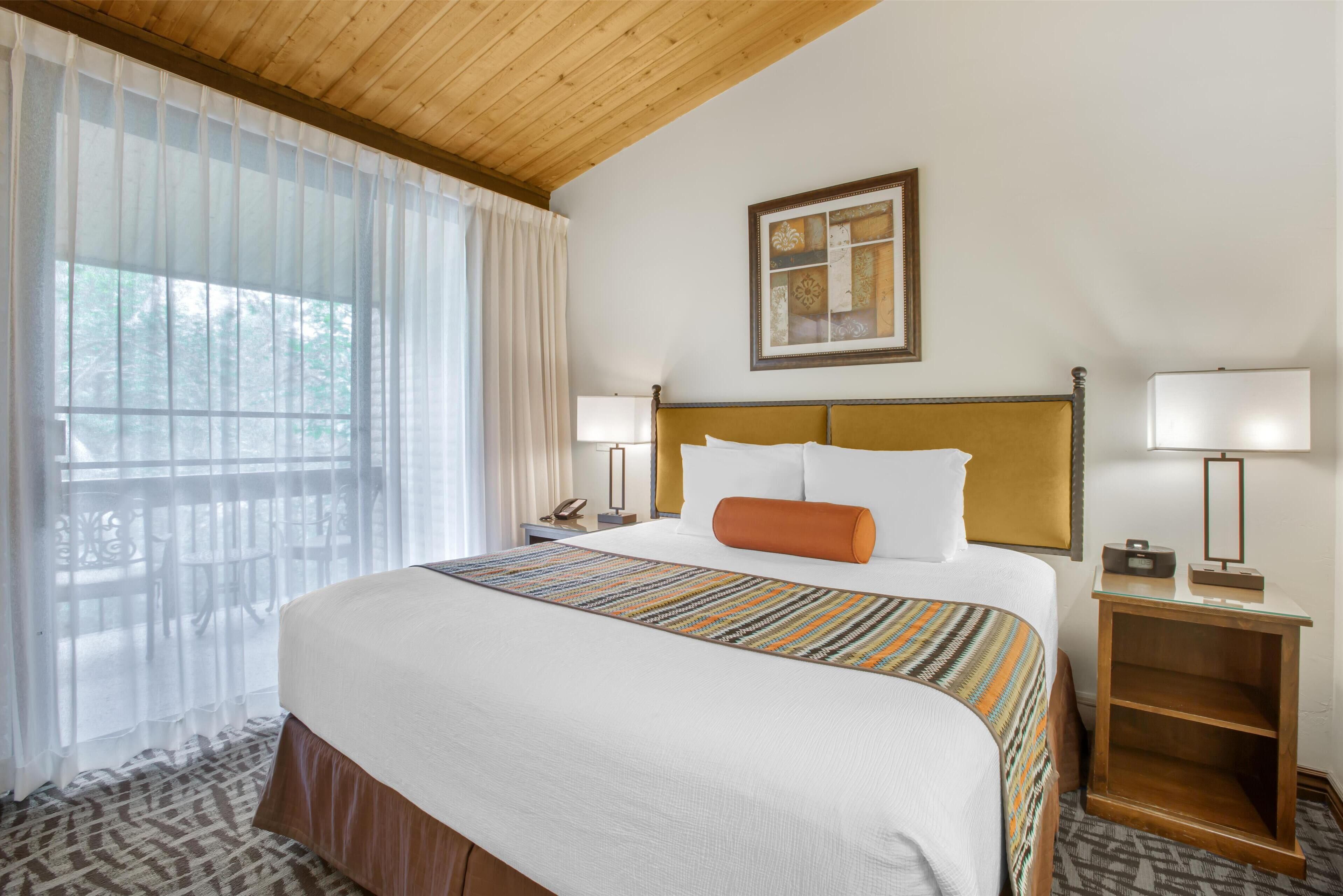 A hotel room with a large bed, a wooden headboard, a bedside table with a lamp, and a window with sheer curtains.