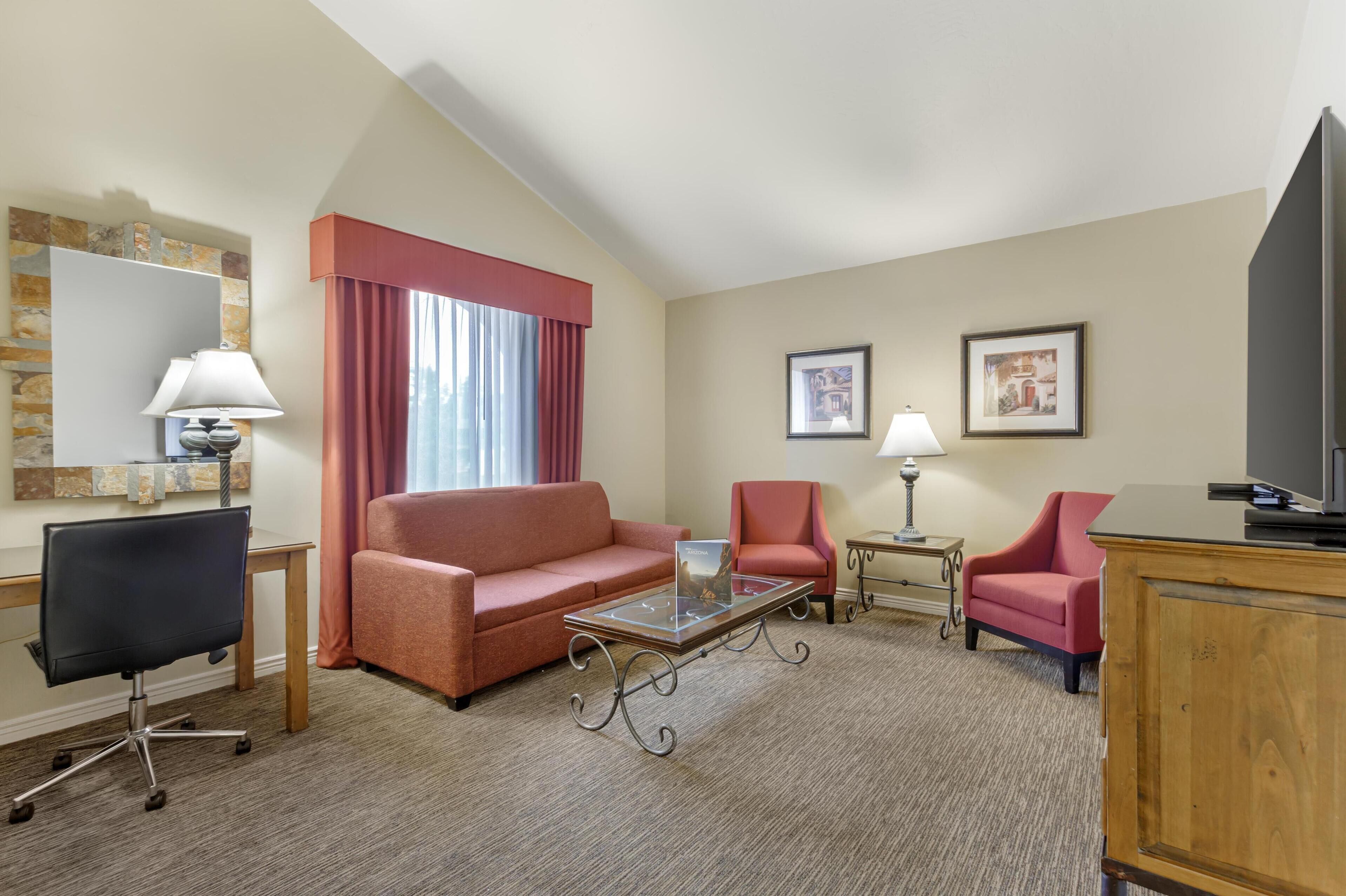 A hotel room with a sofa, two armchairs, a glass coffee table, a desk with a chair, a TV, and two lamps.