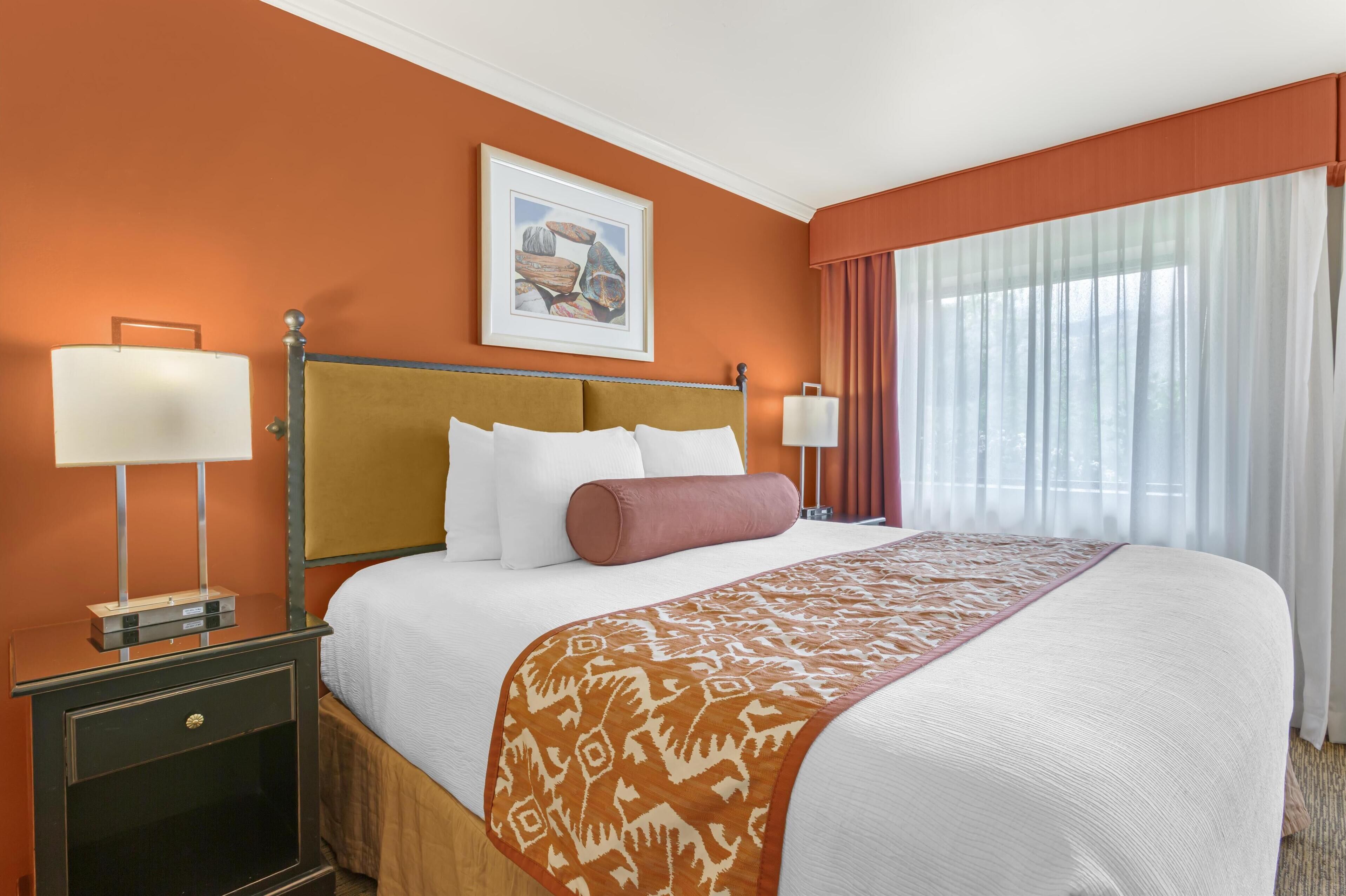 Une chambre d’hôtel avec un grand lit, deux tables de chevet avec des lampes, un tableau encadré au mur et une fenêtre avec des rideaux.