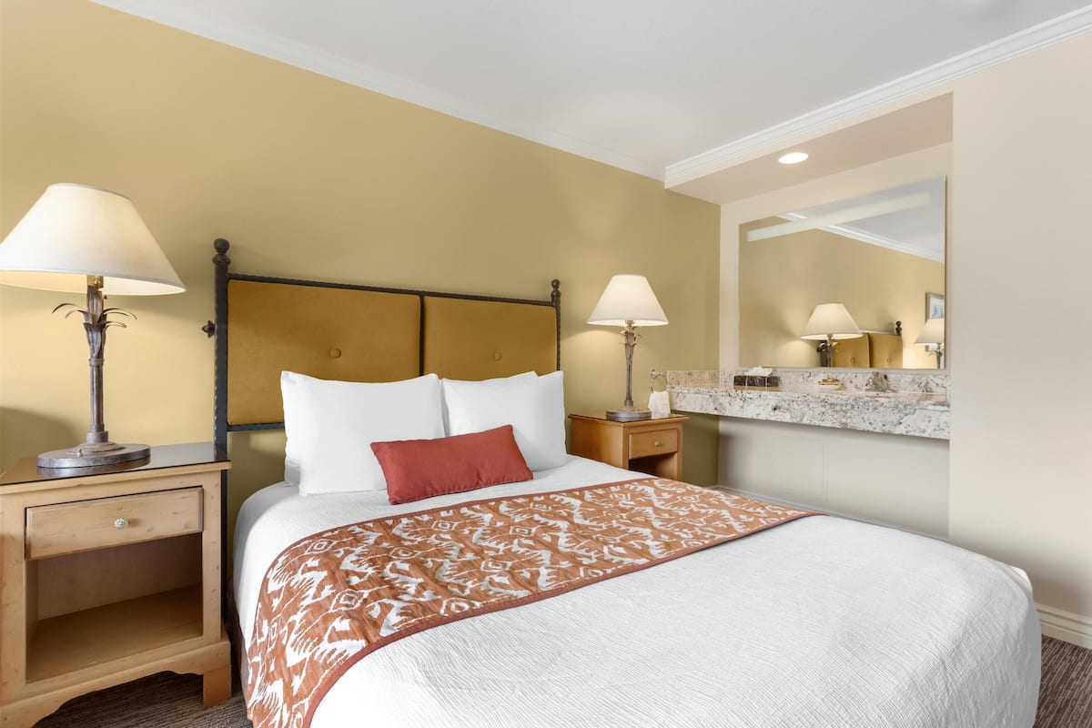Une chambre d’hôtel comprenant un lit, deux tables de chevet avec des lampes, une petite table de nuit avec un tiroir, un miroir encadré de marbre et une étagère murale agrémentée d’objets décoratifs.