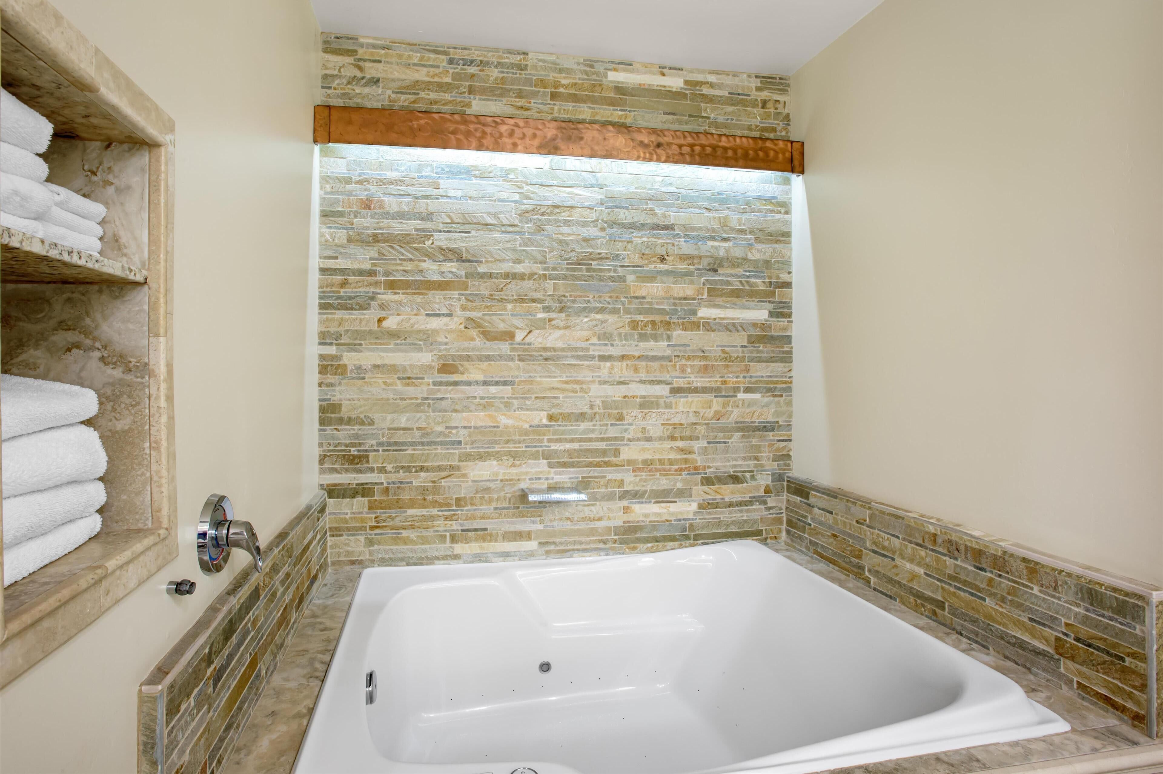 A bathroom with a large bathtub, stone mosaic wall, and a shelf with folded towels.