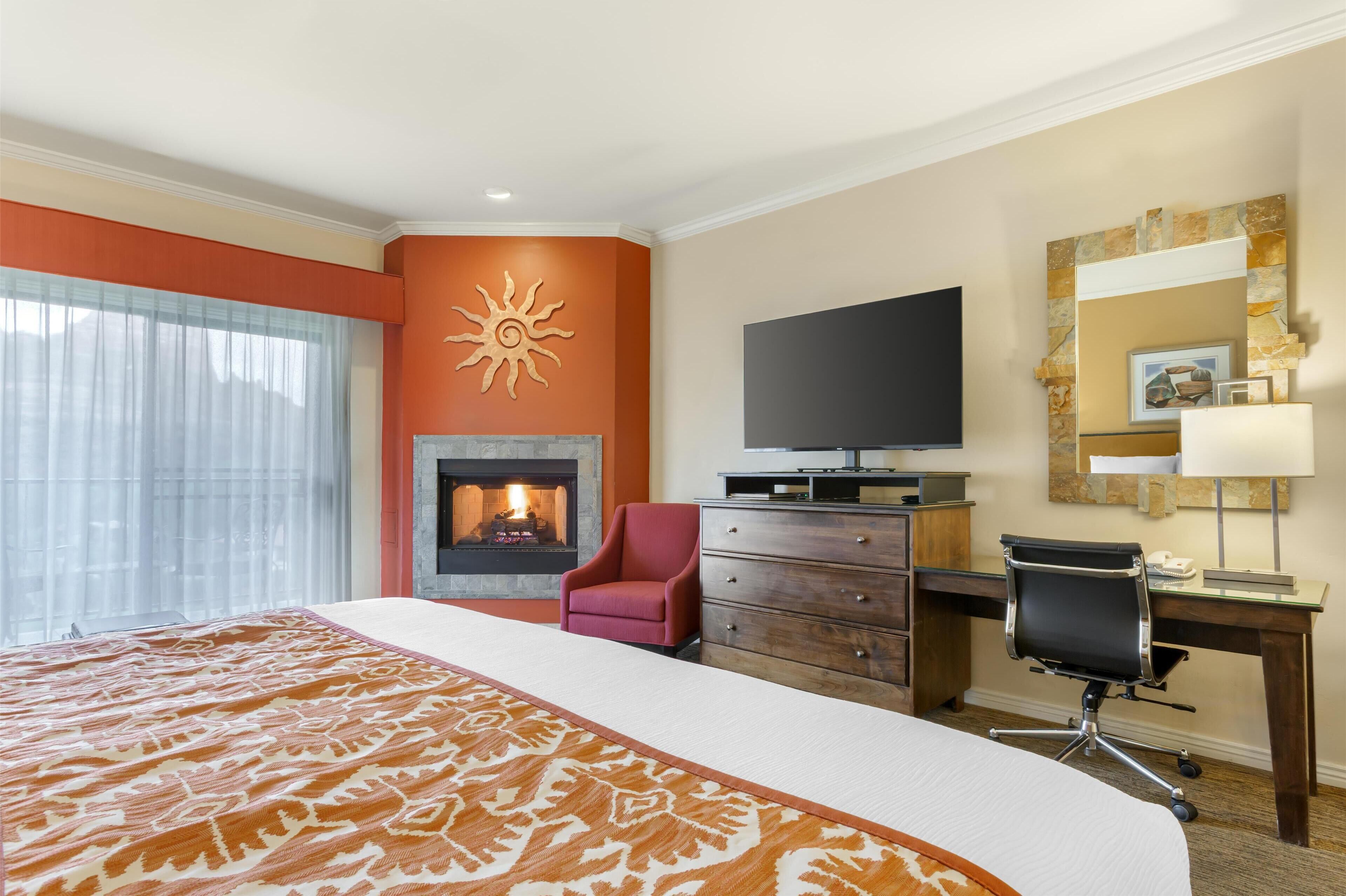 A hotel room with a large bed, a fireplace, a television, a desk with a chair, and a window with curtains.