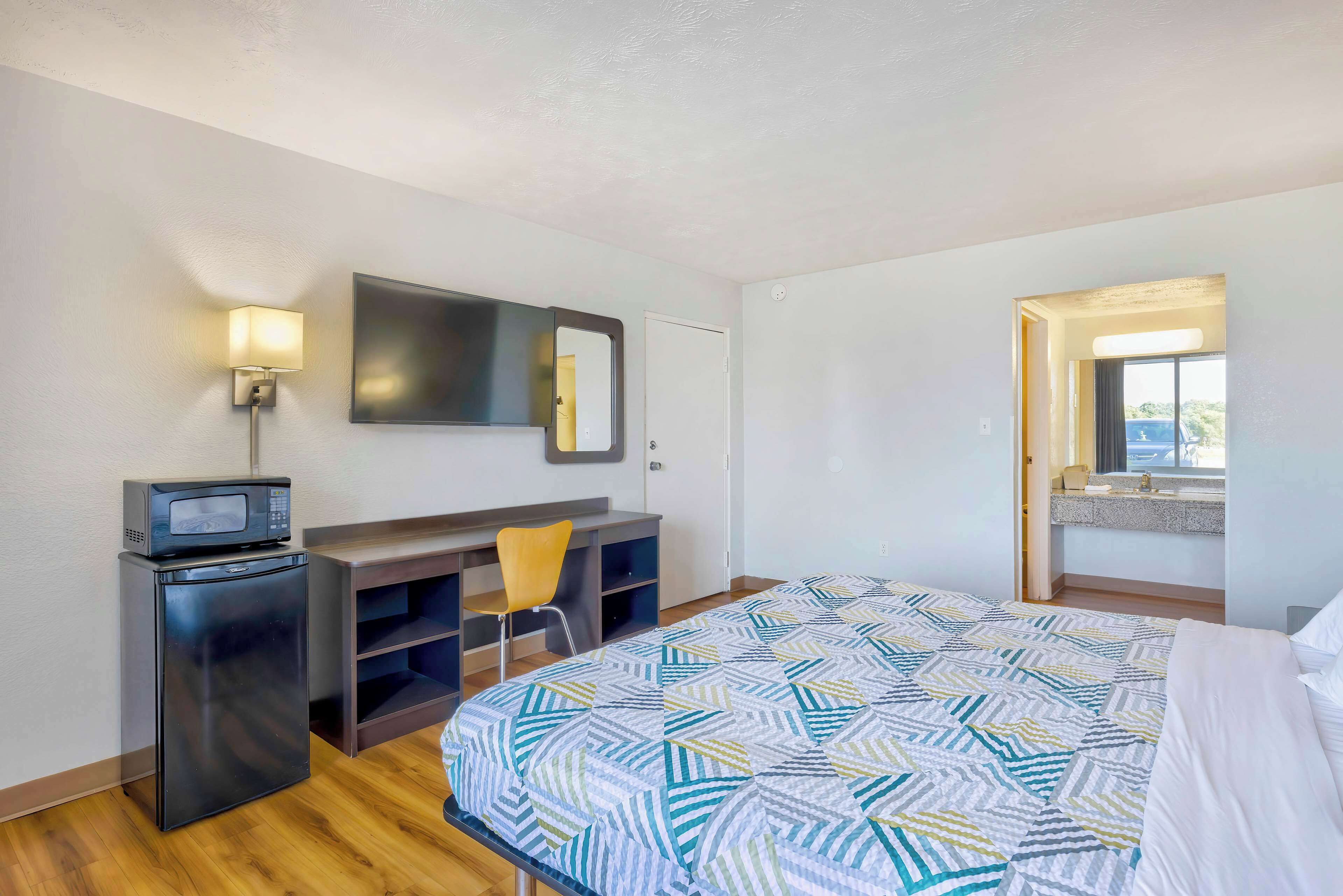 A hotel room with a bed, a desk with a chair, a small refrigerator, a microwave, a wall-mounted TV, and a window with a view.