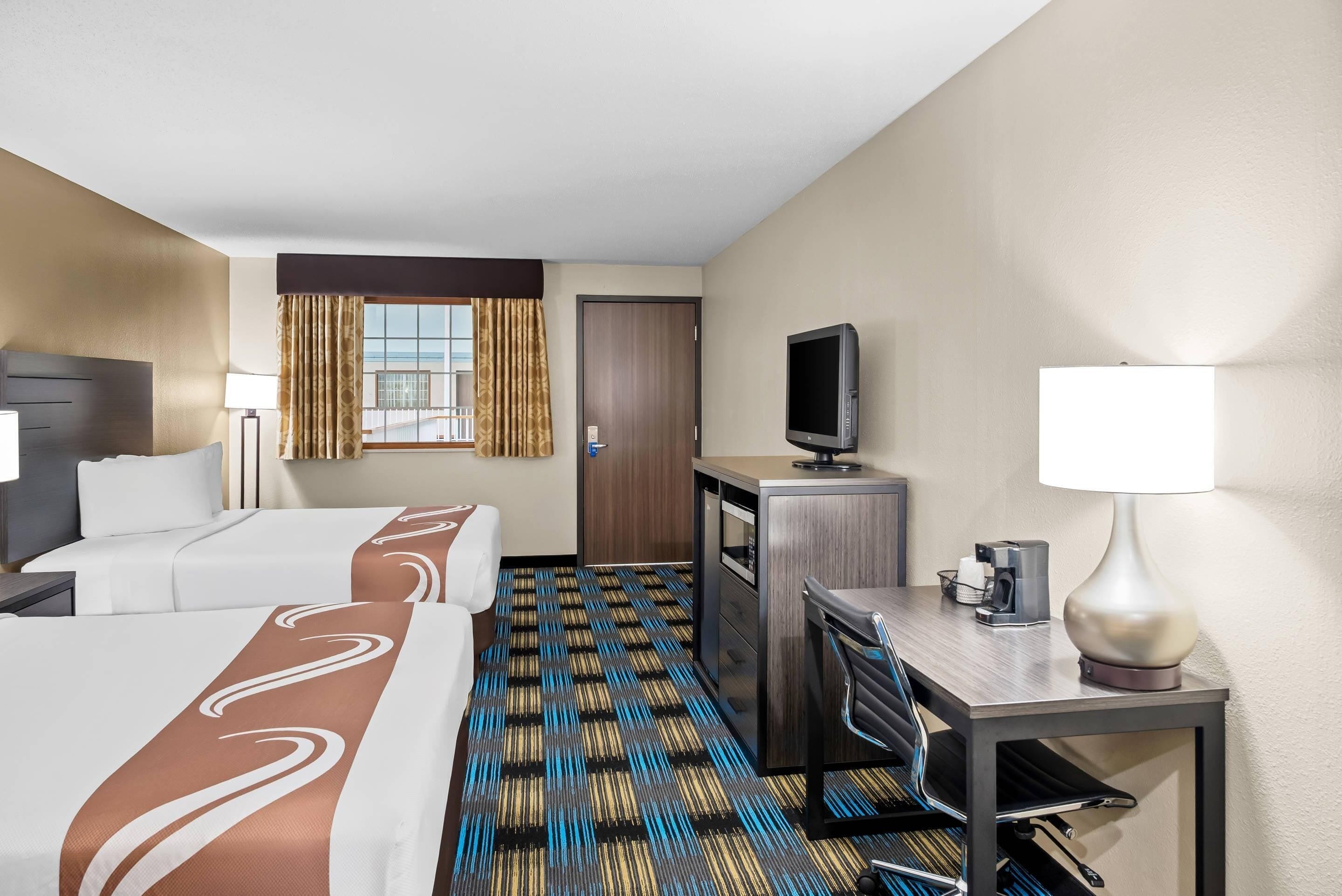 A hotel room with two beds, a desk with a chair, a television, and a lamp.