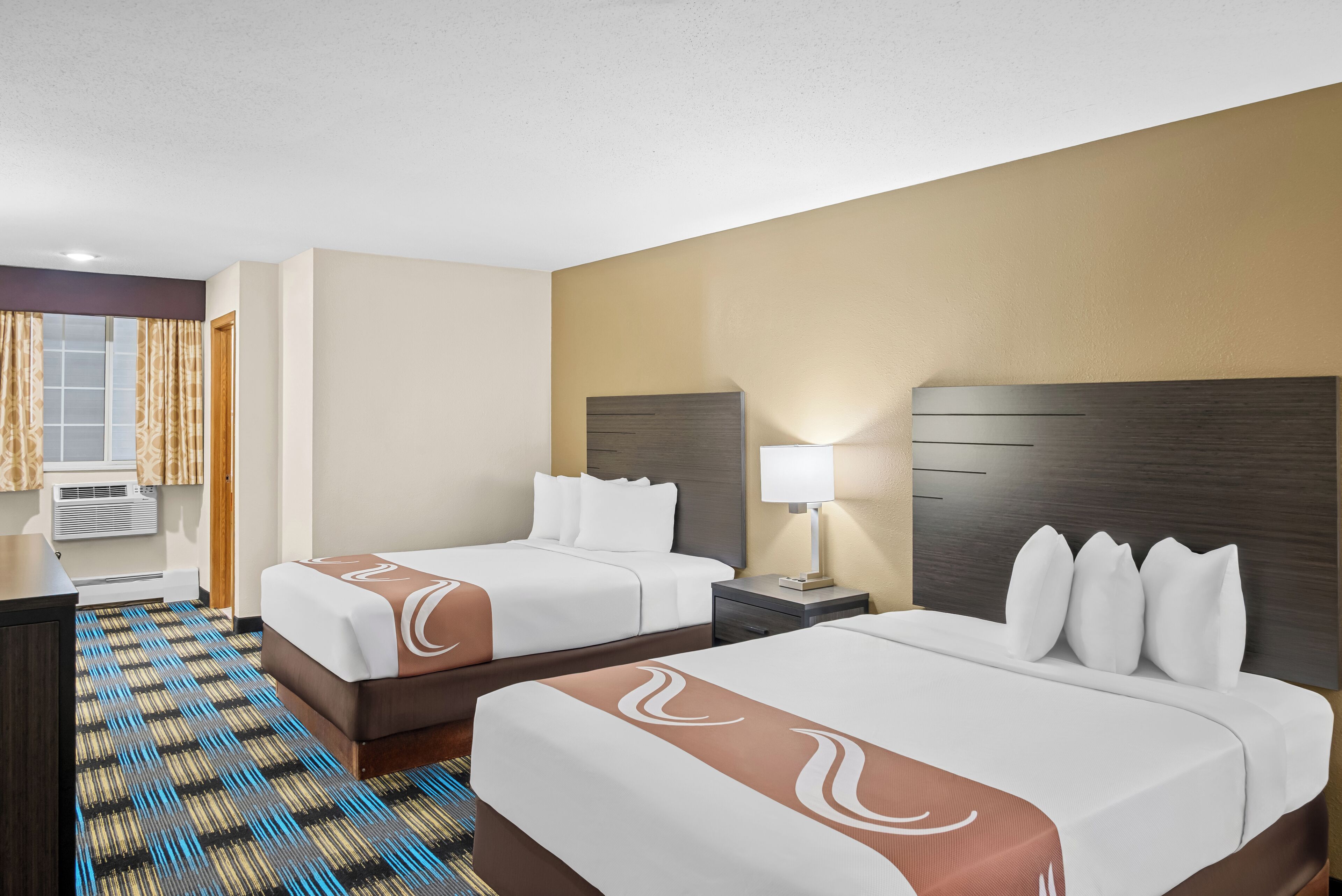 A hotel room with two beds, a desk, a lamp, and a window with curtains.